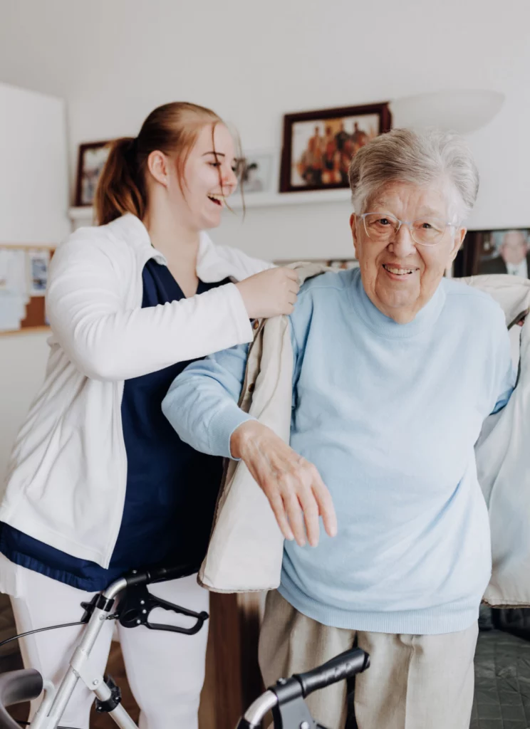 Eine lachende Mitarbeiterin hilft einer lächelnden Seniorin mit Rollator in die Jacke.