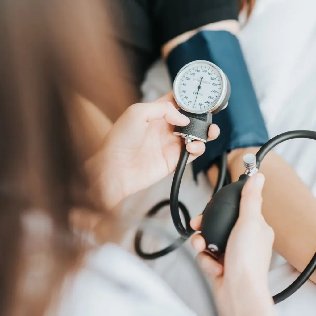 Eine Person misst mit einem analogen Blutdruckmessgerät den Blutdruck einer sitzenden Patientin.