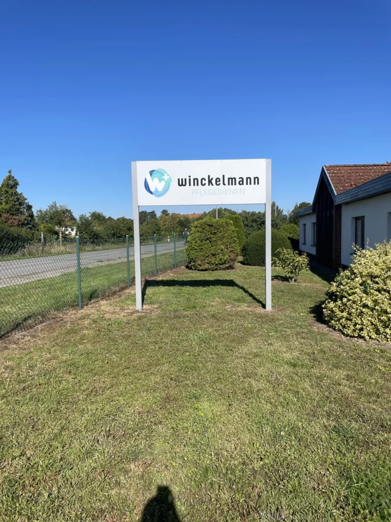 Ein Schild mit dem Logo des Winckelmann Pflegedienstes steht auf einer Rasenfläche vor einem Gebäude.