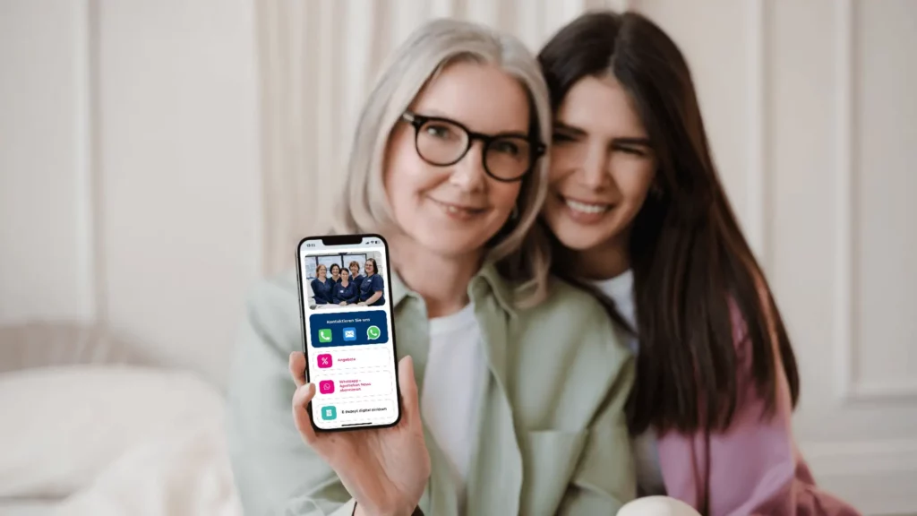 Eine ältere und eine junge Frau sitzen eng beieinander, die ältere hält ein Smartphone. Das Smartphone zeigt die digitale Kundenkarte mit Vorteilen und Funktionen.