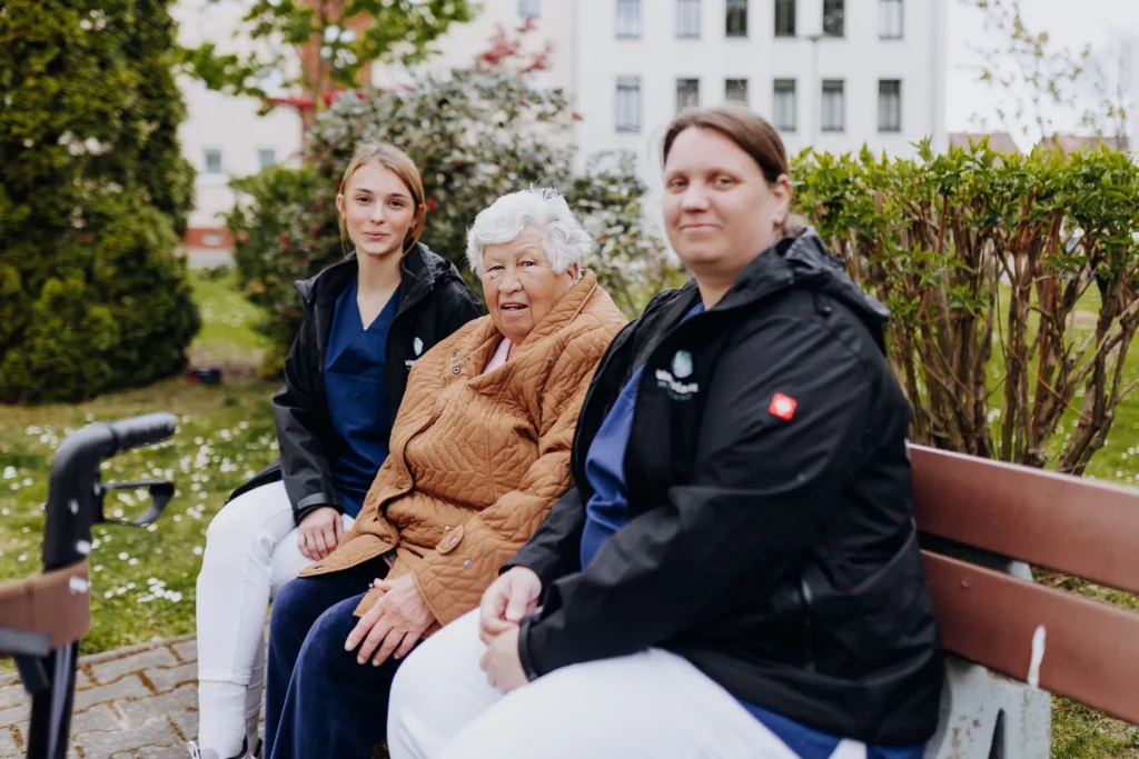 Zwei Mitarbeiterinnen in Dienstjacken sitzen mit einer Seniorin auf einer Parkbank, vorne steht ein Rollator.