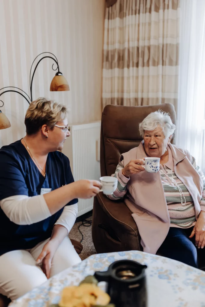 Eine Mitarbeiterin und eine Seniorin im Sessel stoßen im Wohnzimmer mit Kaffeetassen an.