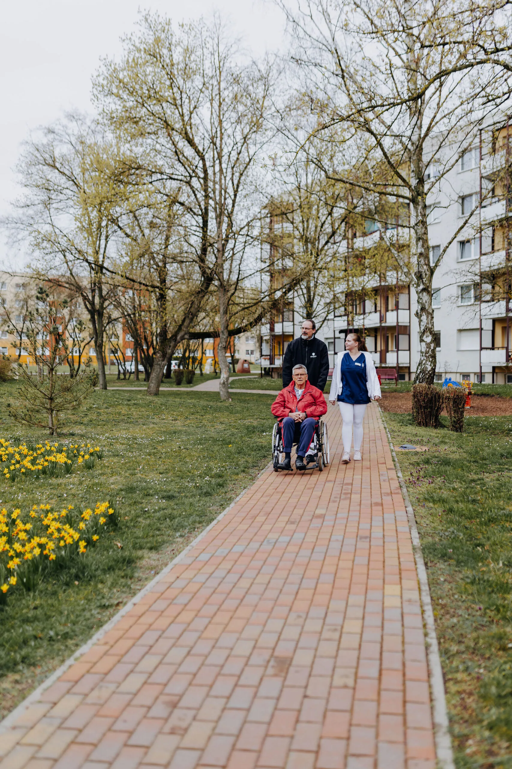 Ein Mitarbeiter schiebt einen Senior im Rollstuhl über einen Gehweg, eine Mitarbeiterin geht nebenher.