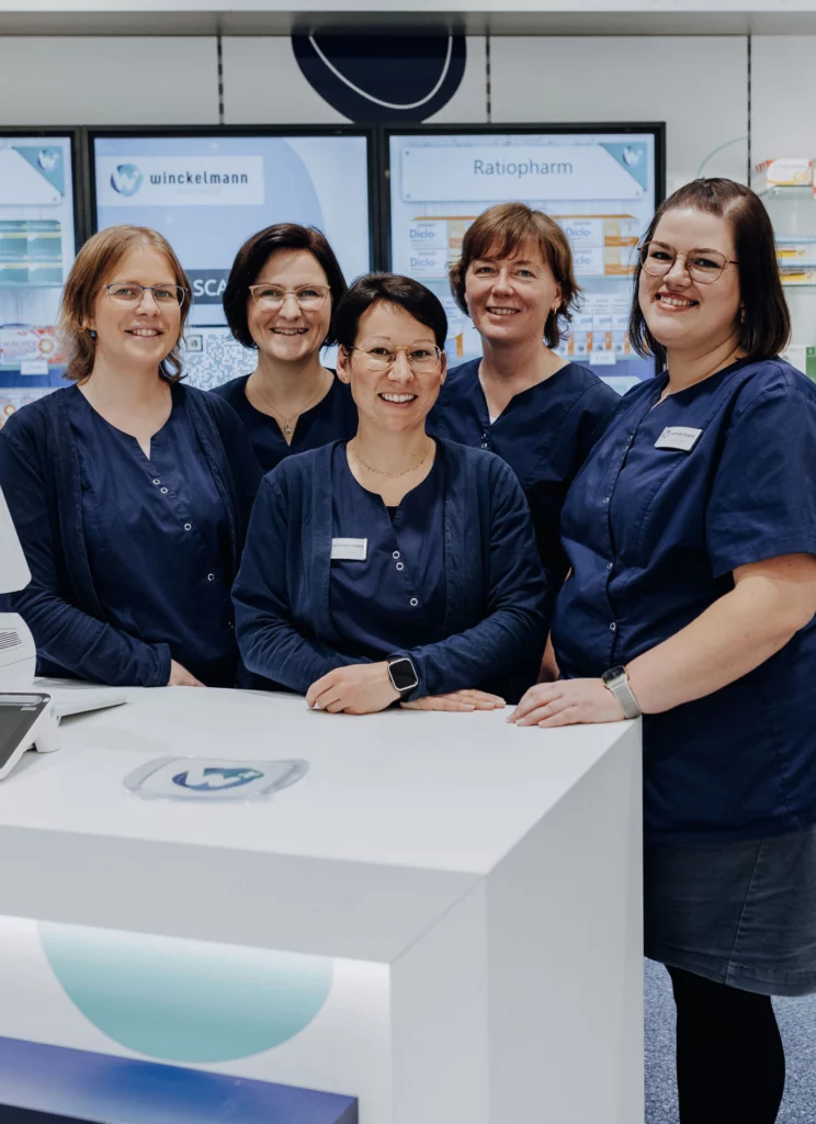 Fünf lächelnde Mitarbeiterinnen der Winckelmann Apotheke stehen gemeinsam an der Beratungstheke.
