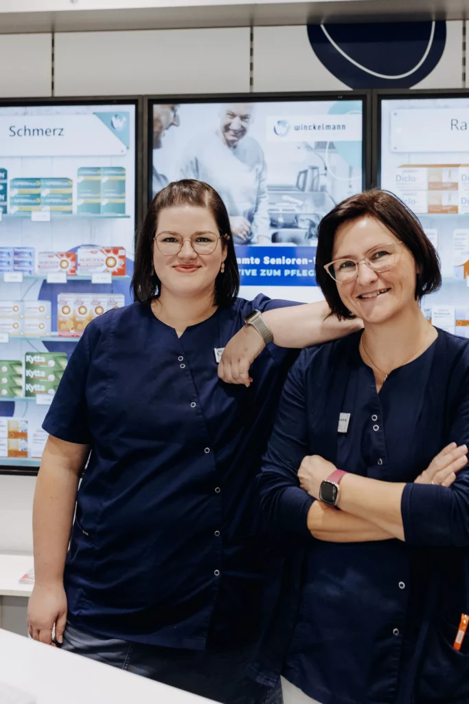 Zwei Mitarbeiterinnen der Winckelmann Apotheke stehen lächelnd und vertraut nebeneinander im Verkaufsraum.
