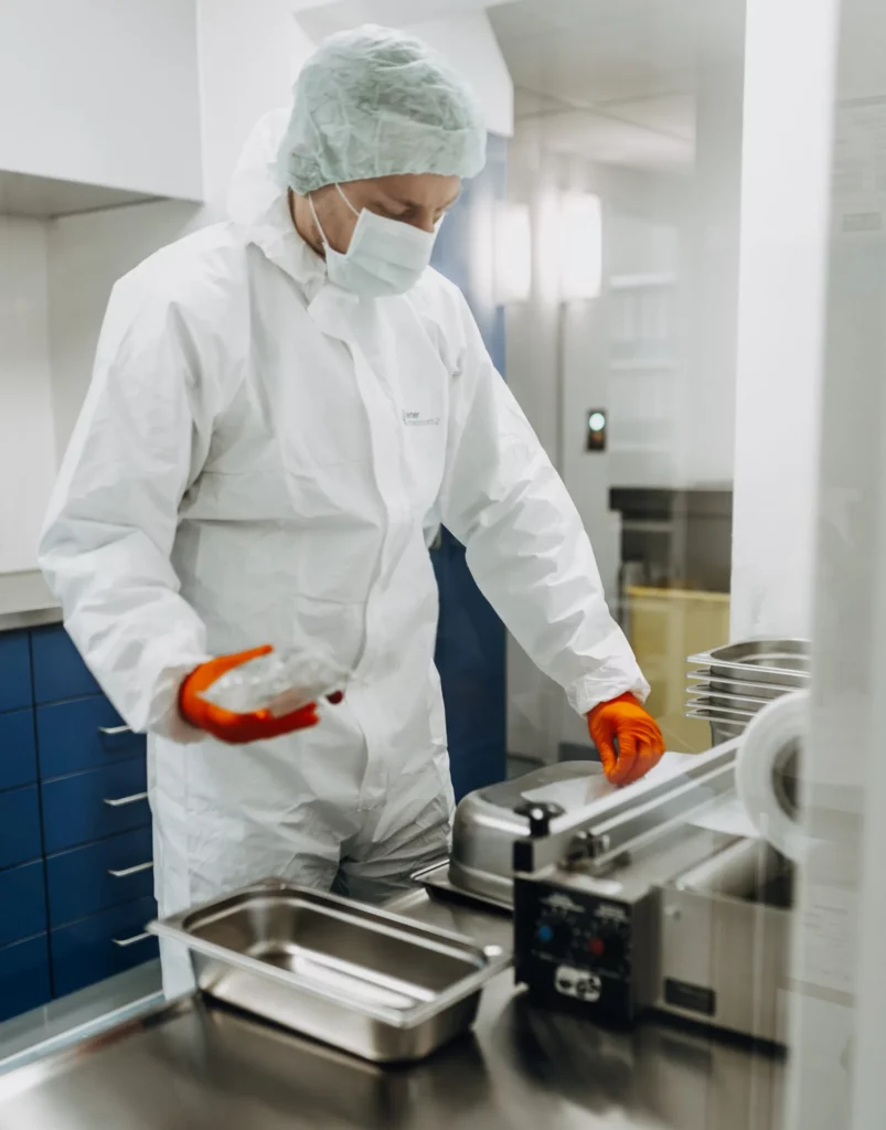 Ein Mitarbeiter in weißer Schutzkleidung, Haube und Mundschutz bedient eine Versiegelungsmaschine in der Rezeptur.
