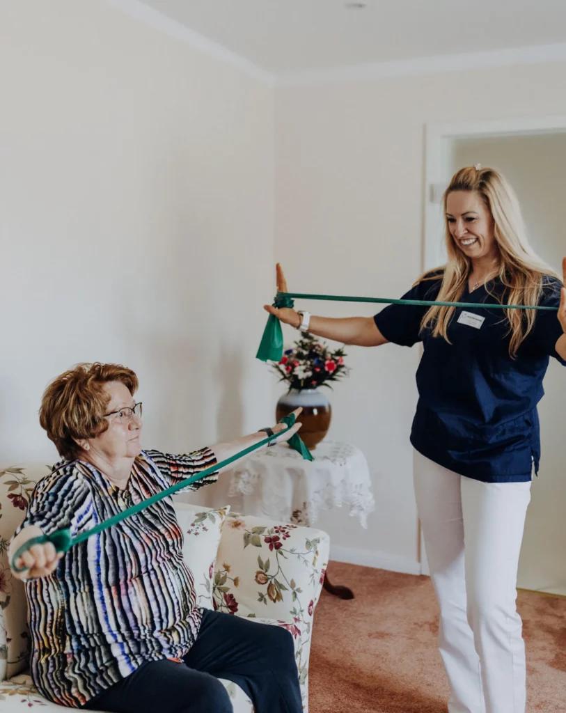 Eine Seniorin sitzt auf einem geblümten Sofa und zieht ein grünes Theraband auseinander, eine Physiotherapeutin macht die Übung stehend vor.