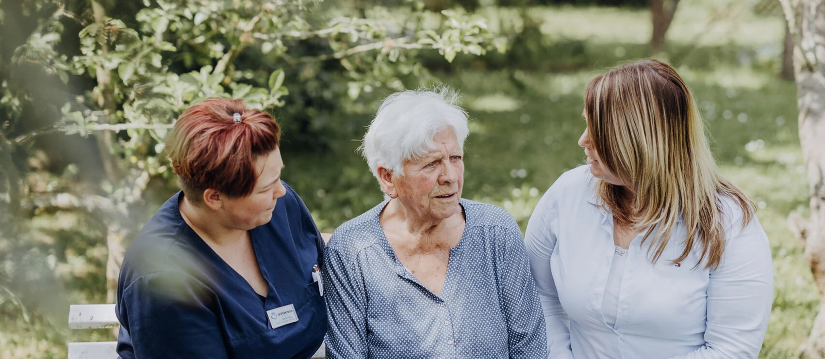 Eine Seniorin sitzt zwischen zwei Mitarbeiterinnen auf einer Gartenbank, beide wenden sich ihr im Gespräch zu.