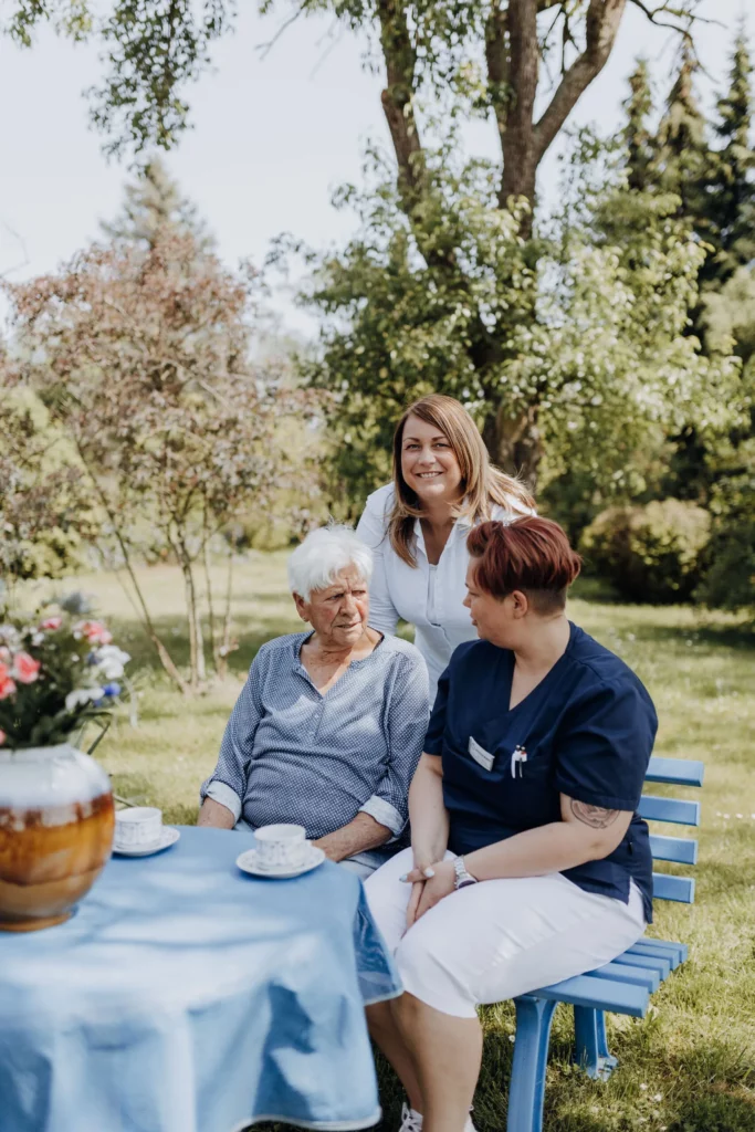 Eine Seniorin und eine Mitarbeiterin sitzen an einem gedeckten Gartentisch, dahinter steht lächelnd eine Frau in weißer Bluse.