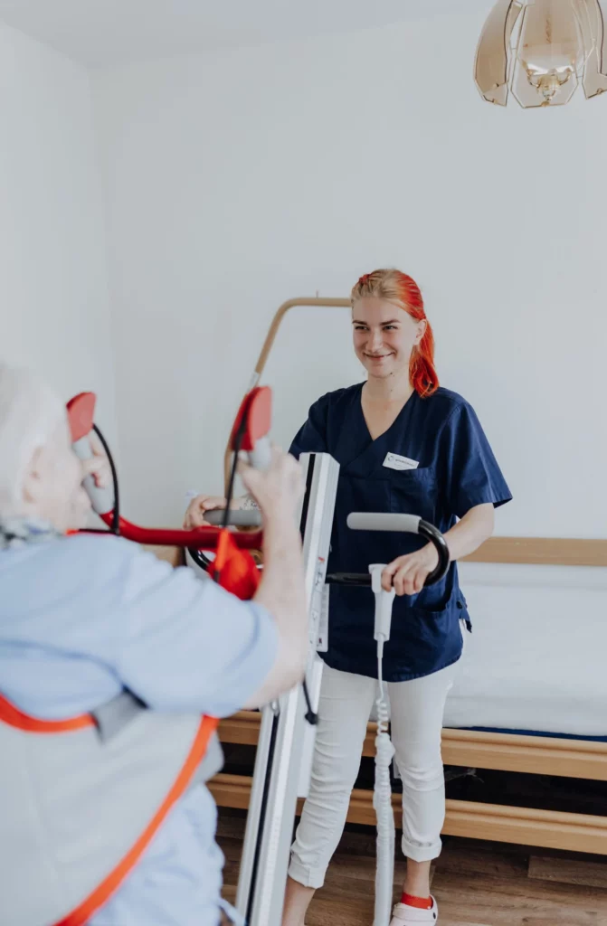 Eine lächelnde Mitarbeiterin bedient einen Patientenlifter, an dessen Griffen sich eine Seniorin festhält.