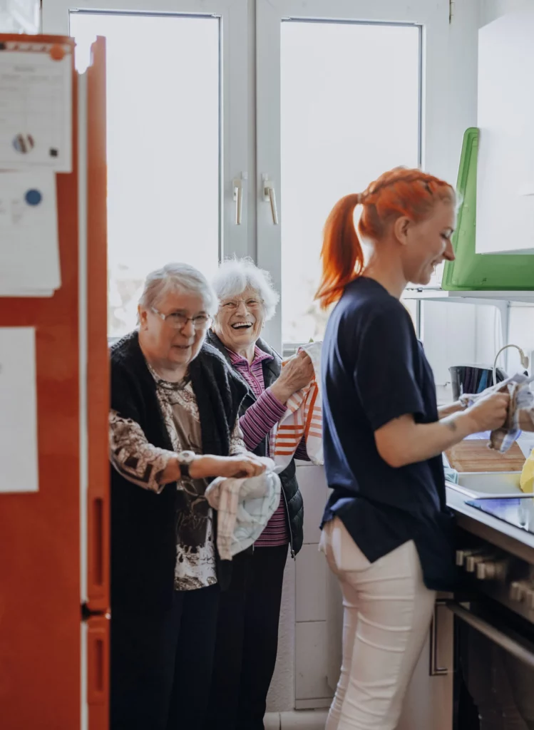 Zwei lachende Seniorinnen trocknen gemeinsam mit einer Mitarbeiterin Geschirr in der Küche ab.