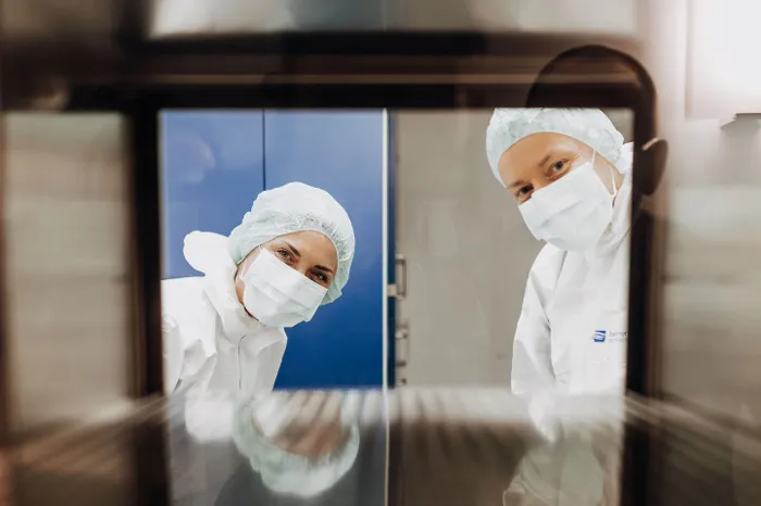 Zwei Personen in Schutzkleidung mit Haube und Mundschutz blicken durch eine Glasscheibe des Reinraums.