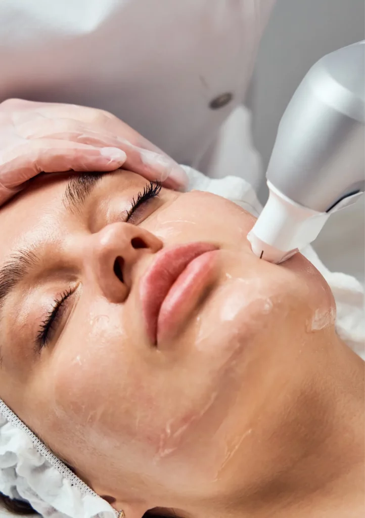 Eine Kosmetikerin mit Handschuhen führt ein Ultraschallgerät über das mit Gel bedeckte Gesicht einer liegenden Frau.