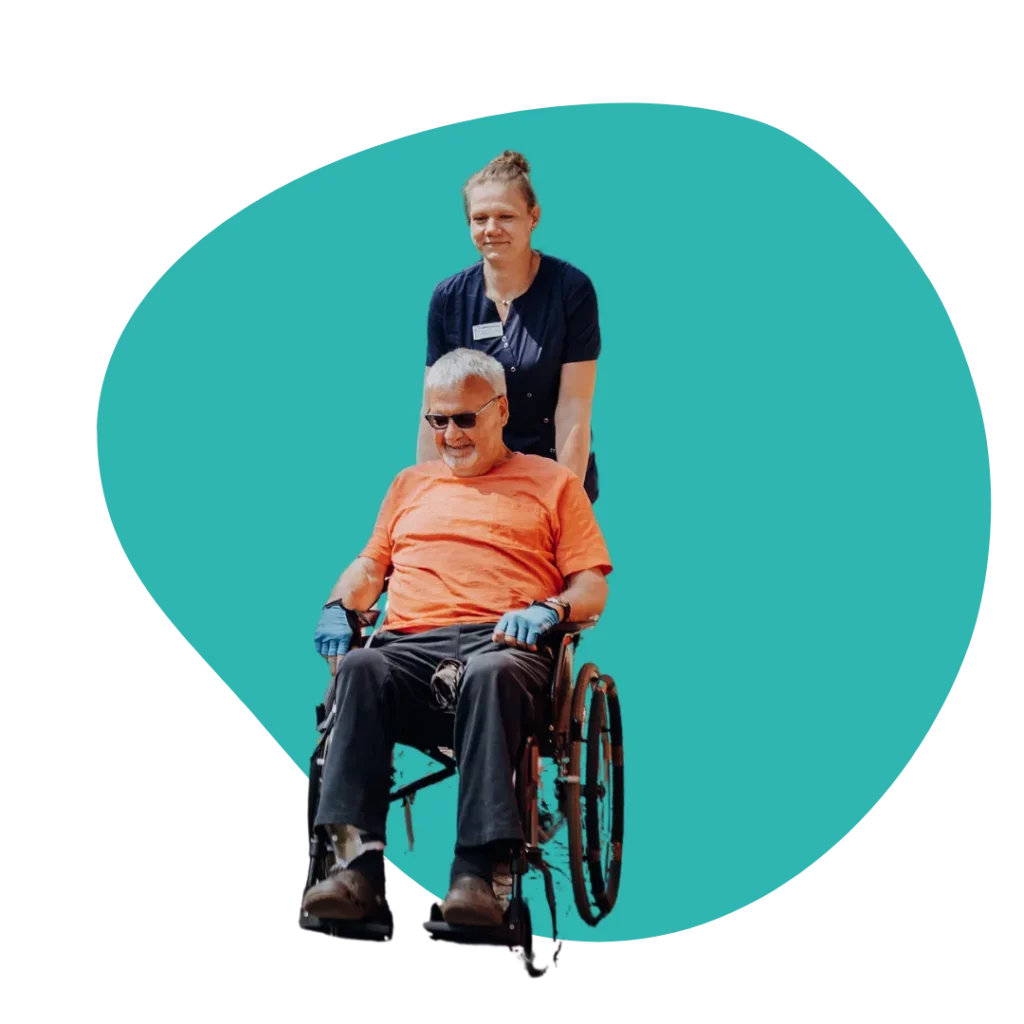 Eine Mitarbeiterin schiebt einen lächelnden Senior mit Sonnenbrille und orangem T-Shirt im Rollstuhl.
