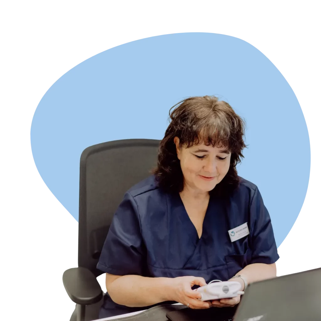Eine Fachkrankenschwester sitzt am Schreibtisch vor einem Laptop und hält ein kleines Gerät in den Händen.