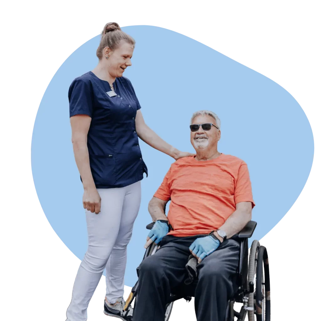 Eine lächelnde Gesundheits- und Krankenpflegerin legt einem Senior im Rollstuhl mit Sonnenbrille die Hand auf die Schulter.