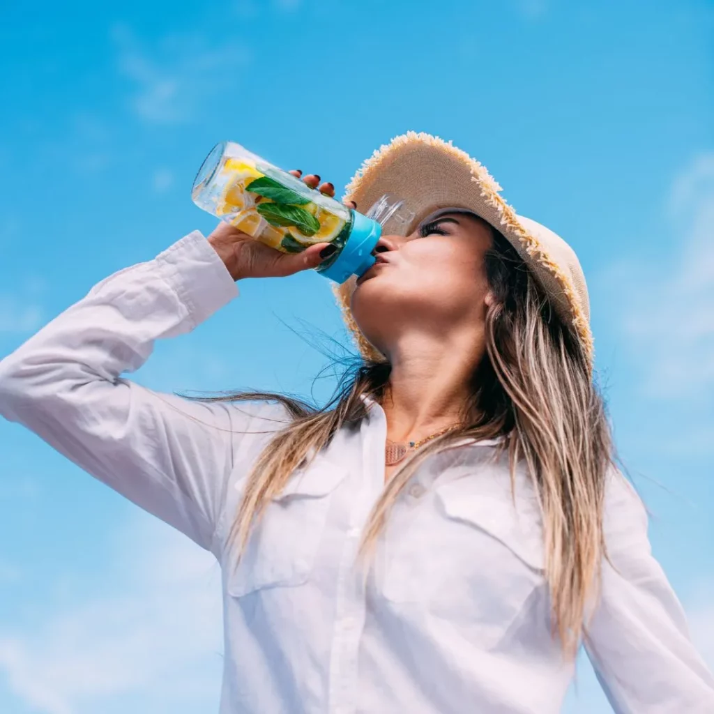 Eine Frau mit Strohhut trinkt vor blauem Himmel aus einer Trinkflasche mit Zitronenscheiben und Minze.