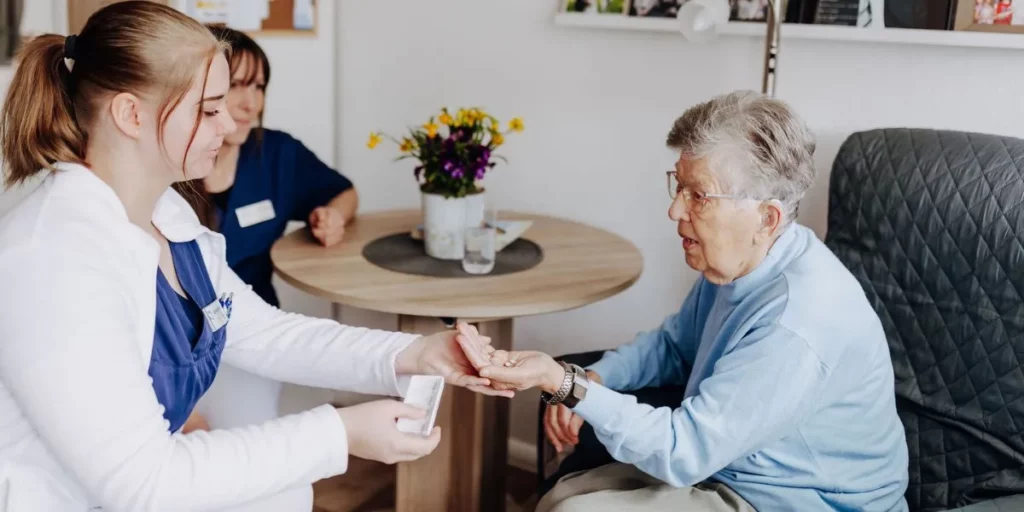 Eine Pflege-Auszubildende reicht einer sitzenden Seniorin Tabletten in die Hand, eine zweite Mitarbeiterin sitzt im Hintergrund am Tisch.