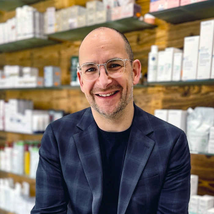 Ein lächelnder Mann im Sakko sitzt vor einem Regal mit Kosmetik- und Pflegeprodukten.