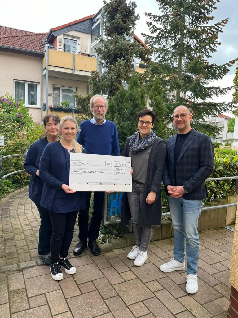 Fünf Personen stehen vor einem Wohnhaus und halten gemeinsam einen Spendenscheck über 2.000 Euro an das Evangelische Hospiz Stendal, datiert auf den 24.04.2024.
