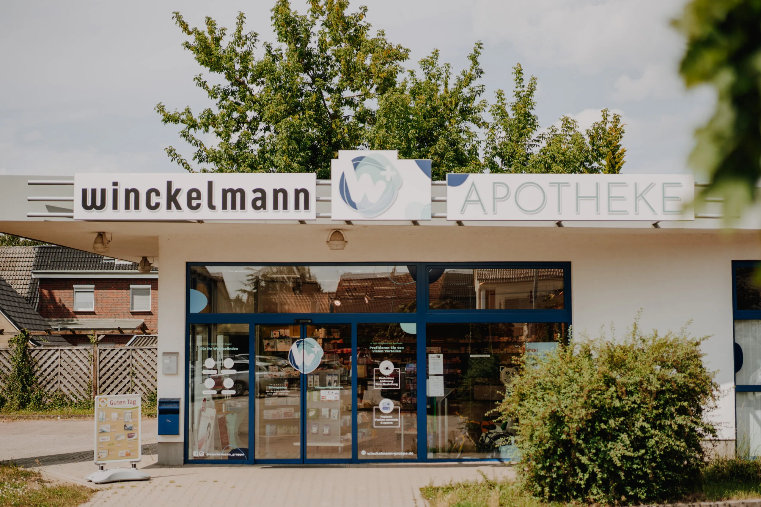 Außenansicht der Winckelmann Apotheke Osterburg, umgeben von Bäumen, mit Info-Aufsteller vor dem Eingang.