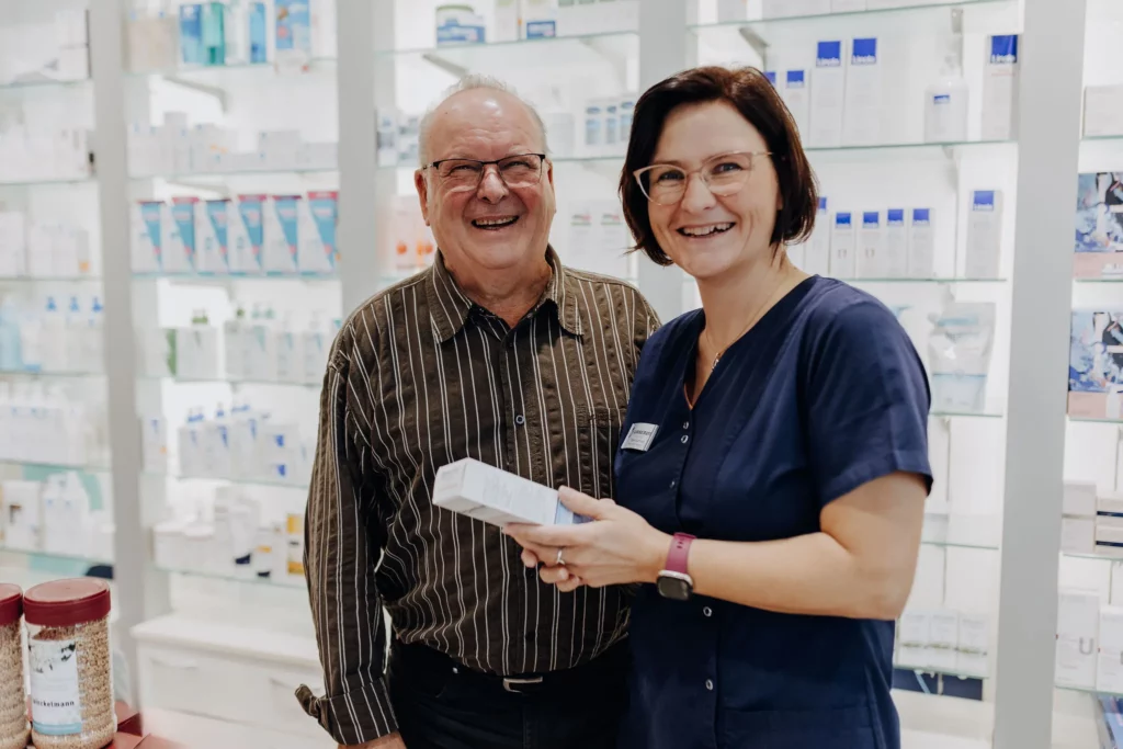 Eine Apothekenmitarbeiterin übergibt lachend eine Medikamentenschachtel an einen ebenfalls lachenden älteren Kunden.