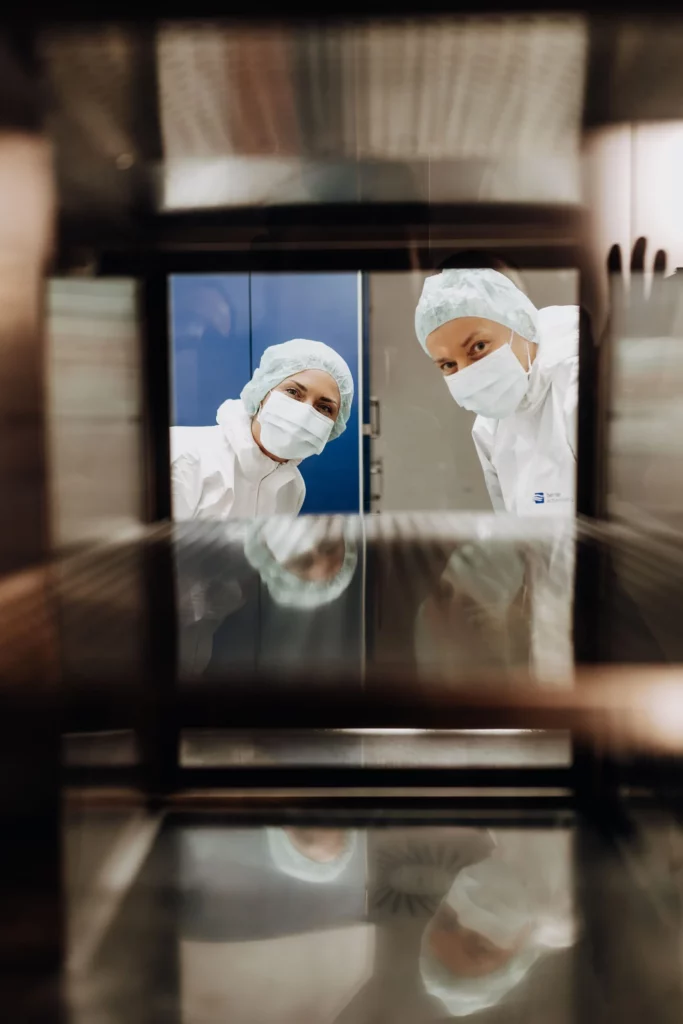Zwei Personen in Schutzkleidung mit Haube und Mundschutz blicken durch eine Glasscheibe des Reinraums.