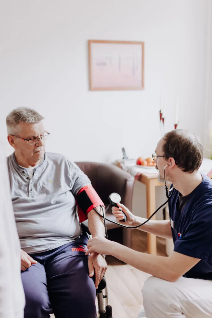 Eine Homecare-Fachkraft mit Stethoskop misst einem Senior im Rollstuhl mit einer roten Manschette den Blutdruck.