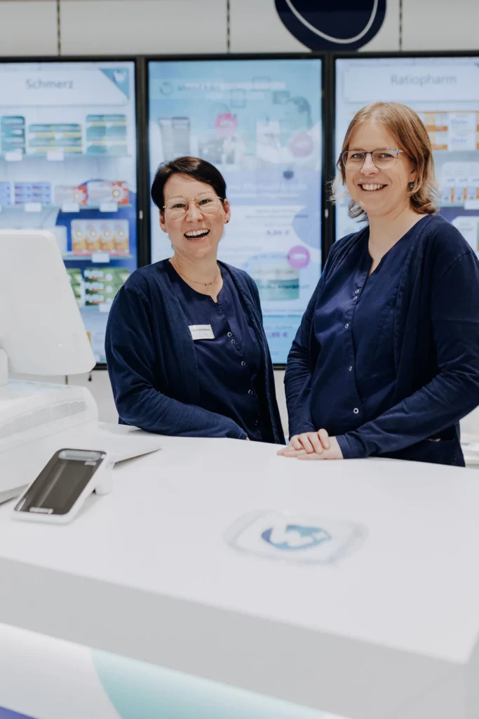 Zwei lächelnde Mitarbeiterinnen stehen nebeneinander an der Beratungstheke der Apotheke.