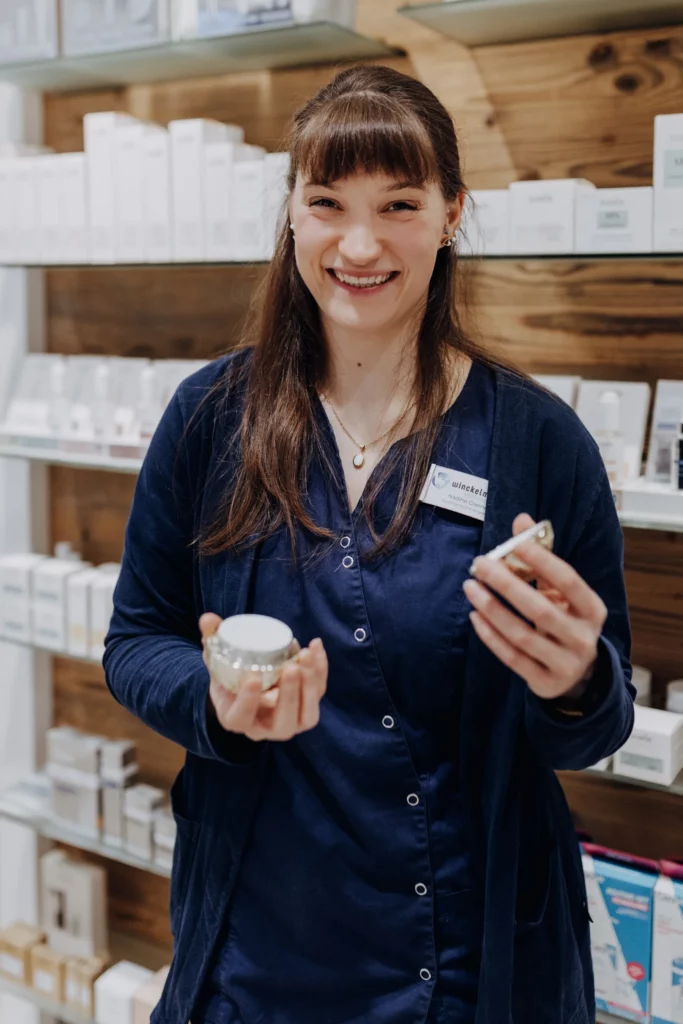 Eine lächelnde Mitarbeiterin hält ein geöffnetes Cremeglas und einen Pinsel in den Händen.