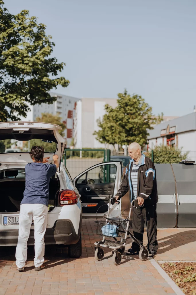 Eine Mitarbeiterin lädt den Kofferraum eines Autos, während ein Mann mit Rollator daneben wartet.