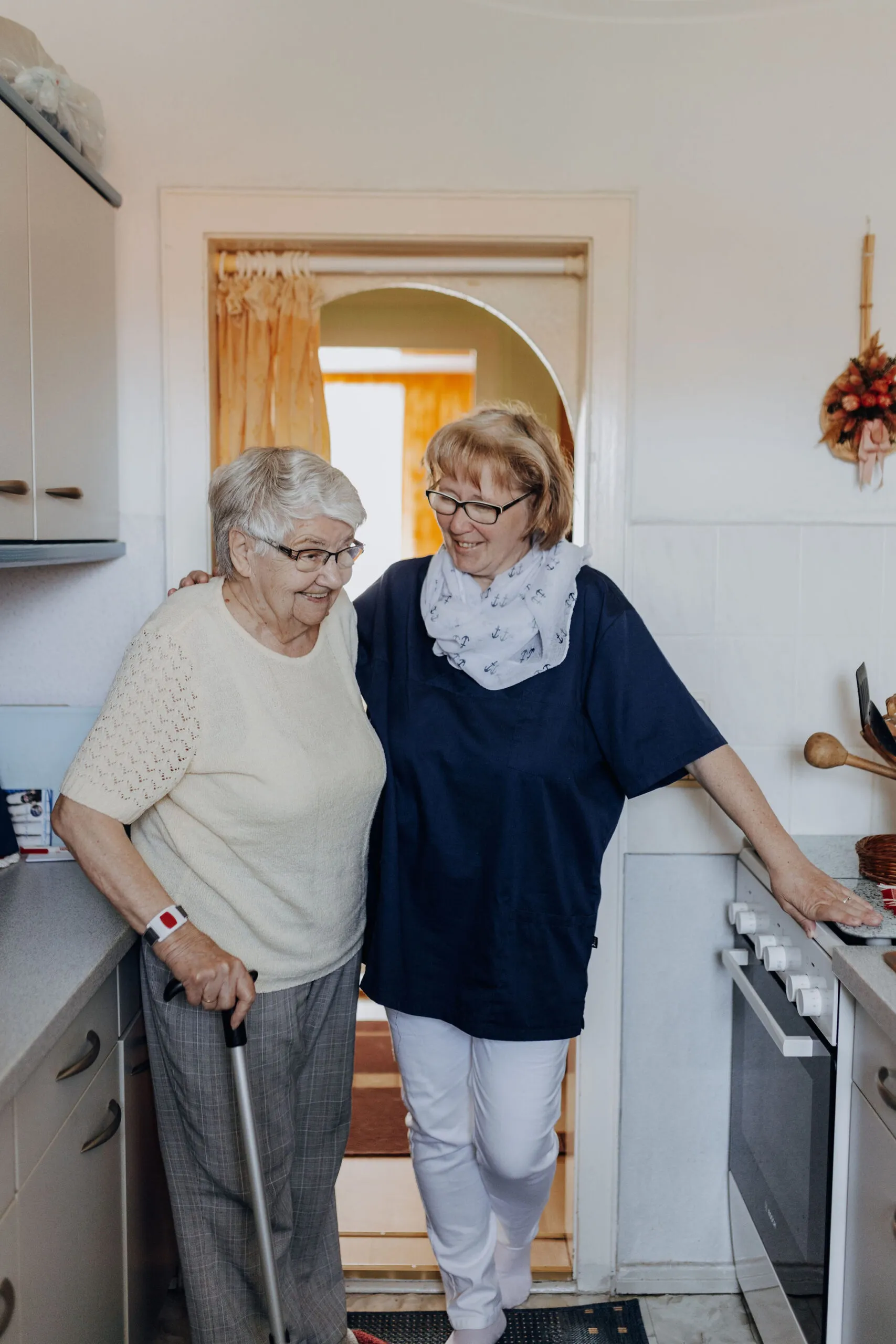 Eine Mitarbeiterin steht stützend neben einer Seniorin mit Gehstock in deren Küche.