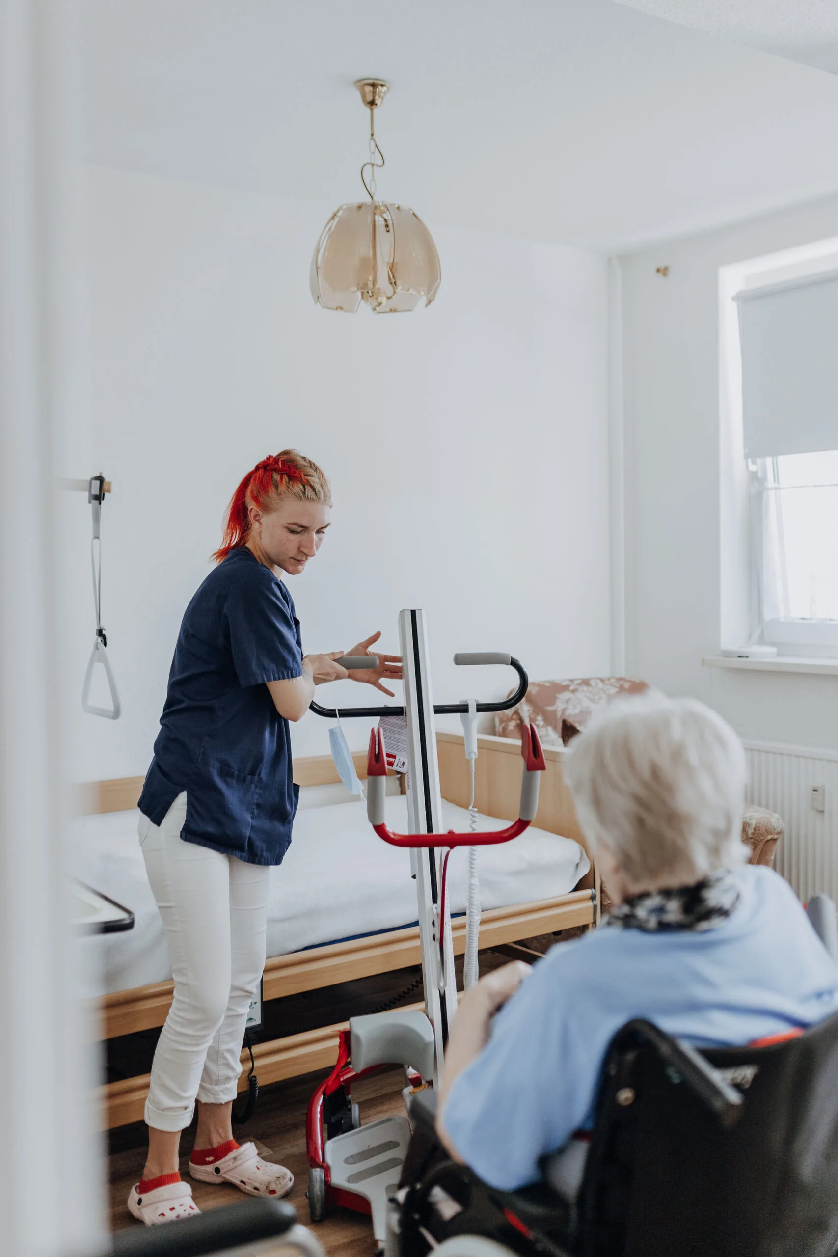 Eine Mitarbeiterin richtet neben einem Pflegebett einen Patientenlifter ein, im Vordergrund sitzt eine Seniorin im Rollstuhl.