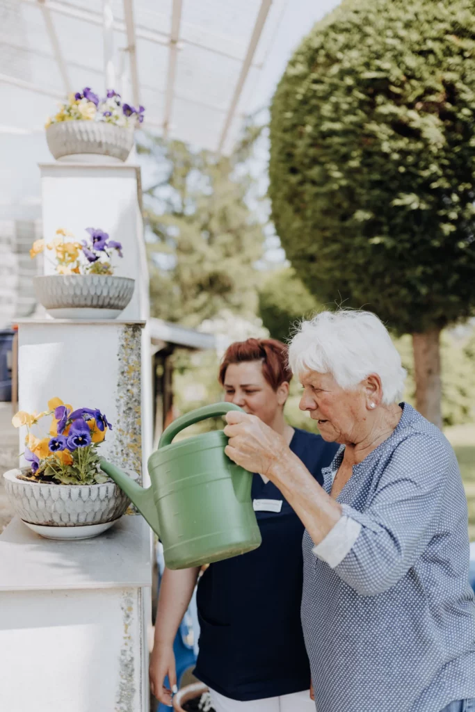 Eine Seniorin gießt mit einer grünen Kanne Blumen, eine Mitarbeiterin steht begleitend daneben.