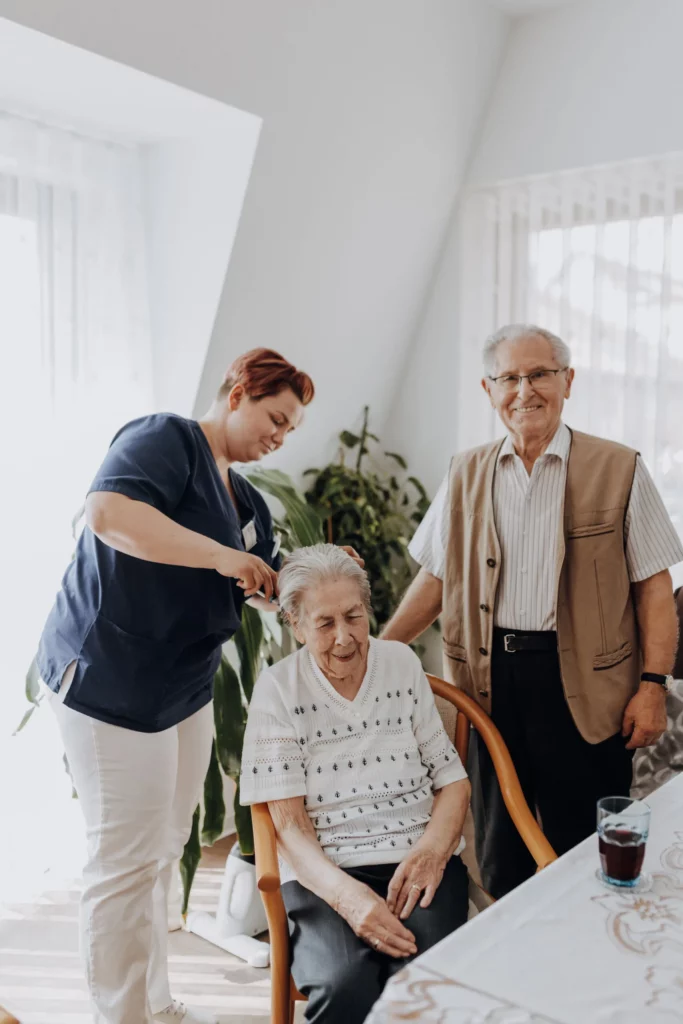 Eine Mitarbeiterin kämmt einer sitzenden Seniorin die Haare, ein älterer Herr steht lächelnd daneben.