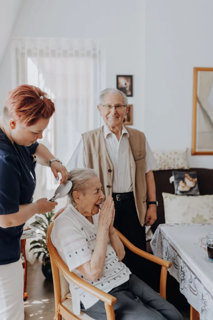 Eine Mitarbeiterin kämmt einer sitzenden Seniorin die Haare, ein älterer Herr steht lächelnd daneben.