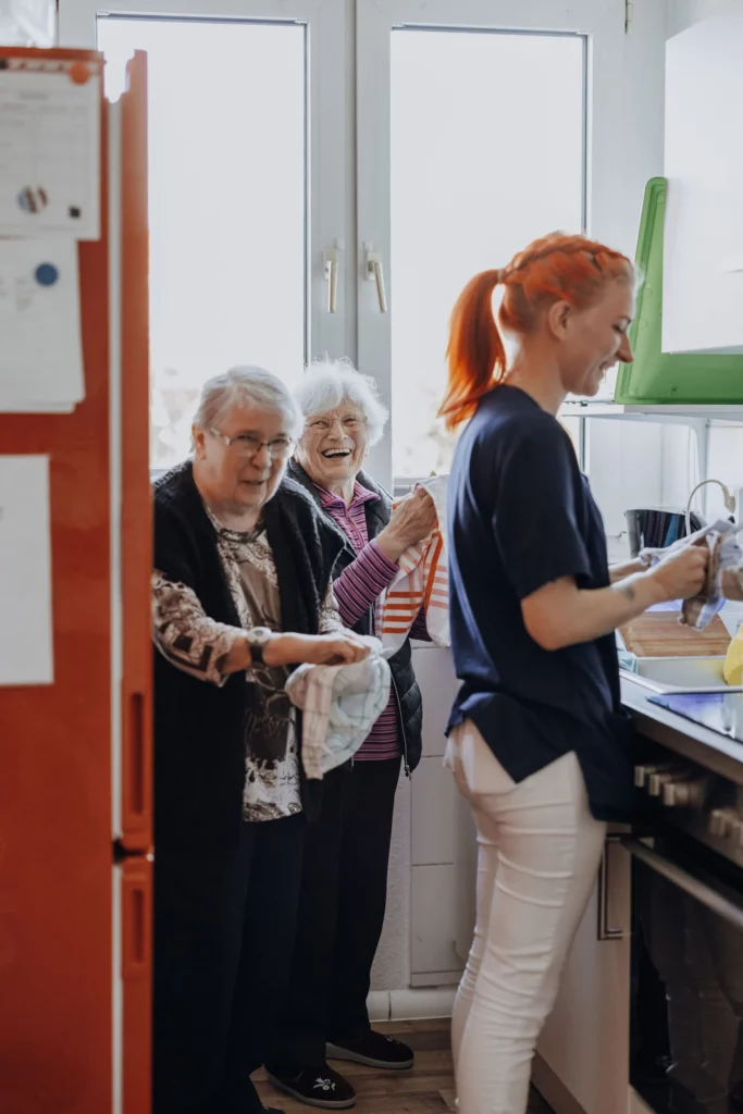 Zwei lachende Seniorinnen trocknen gemeinsam mit einer Mitarbeiterin Geschirr in der Küche ab.