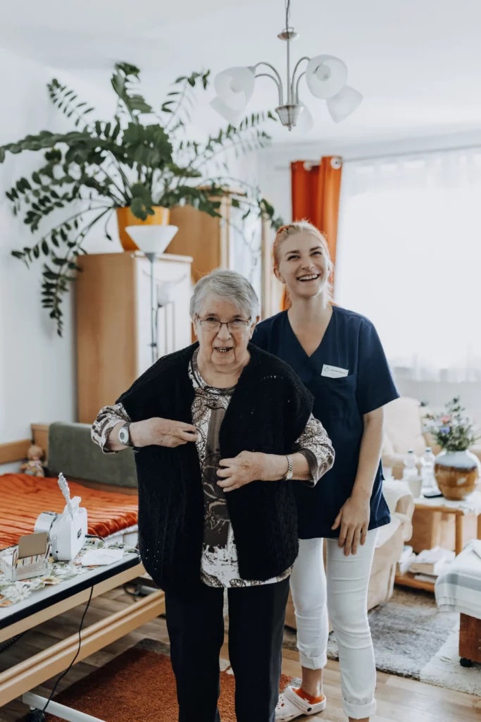 Eine lächelnde Pflegefachkraft steht neben einer Seniorin, die sich eine Strickjacke anzieht.