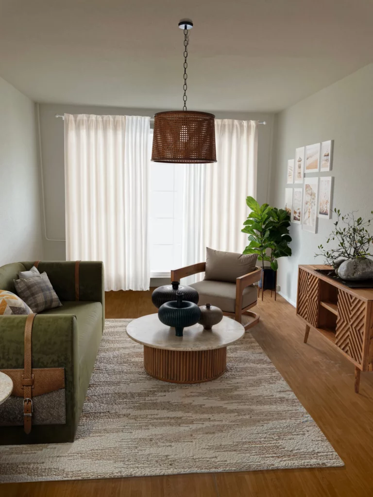 Wohnzimmer mit grünem Sofa, Holzsessel, rundem Couchtisch, Sideboard und Zimmerpflanze vor einem hellen Fenster.