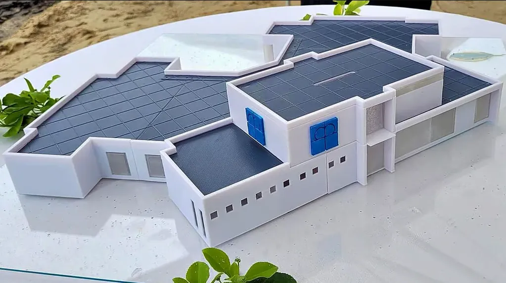 Ein weißes 3D-gedrucktes Architekturmodell zeigt ein verwinkeltes, mehrteiliges Gebäude mit dunklen Dachflächen und blauen Fensterläden, fotografiert auf einem Glastisch im Freien.