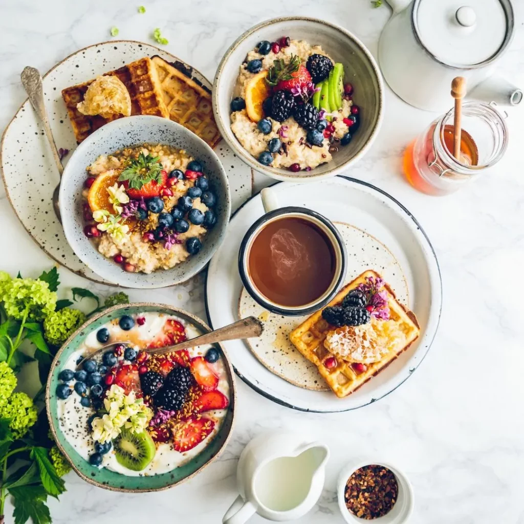 Von oben fotografierter Frühstückstisch mit Porridge-Schalen, einer Joghurtschale mit Beeren und Kiwi, Waffeln, einer Tasse Tee und einem Glas Honig.