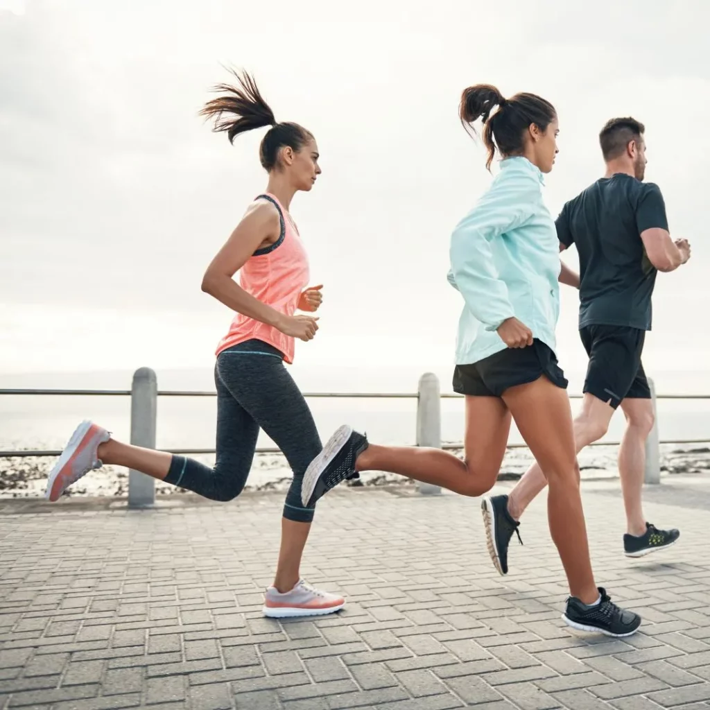 Zwei Frauen und ein Mann joggen in Sportkleidung auf einer gepflasterten Promenade am Meer.