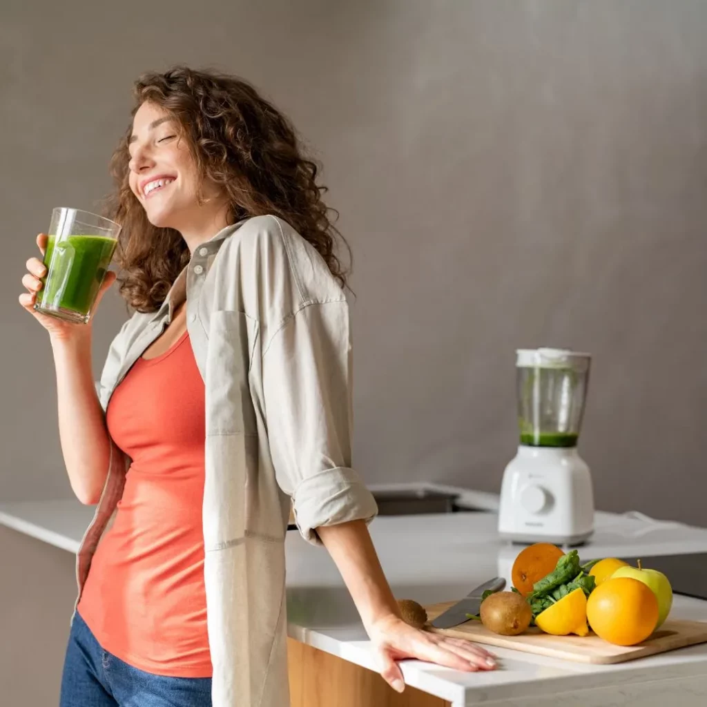 Eine lächelnde Frau mit lockigen Haaren trinkt in der Küche einen grünen Smoothie, neben ihr stehen ein Standmixer und frisches Obst auf einem Holzbrett.
