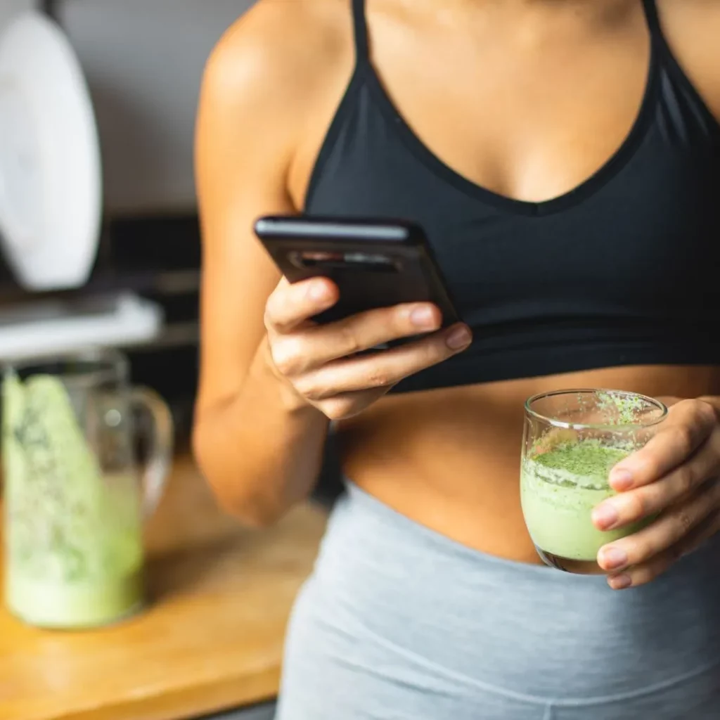 Eine Frau in Sportkleidung hält in der Küche ein Smartphone in der einen und ein Glas grünen Smoothie in der anderen Hand.