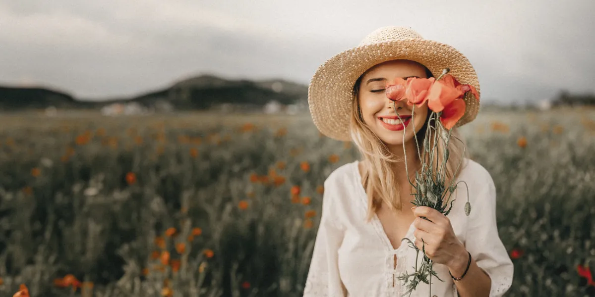 Eine lächelnde Frau mit Strohhut steht in einem Mohnfeld und hält sich einen Strauß Mohnblumen vor das Gesicht.